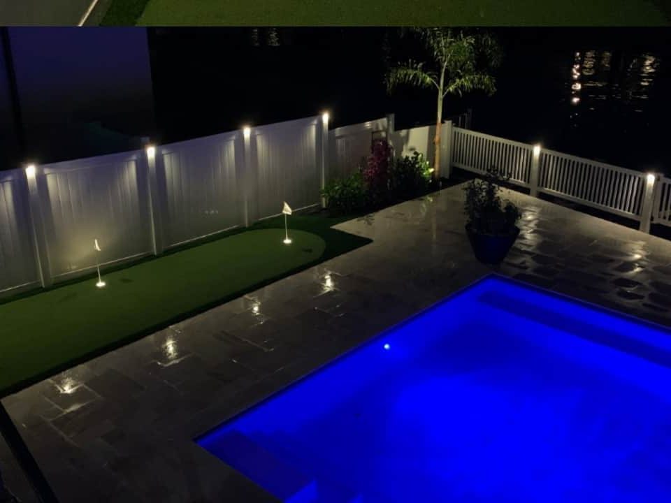 Putting green and pool at night with lighting along a white fence